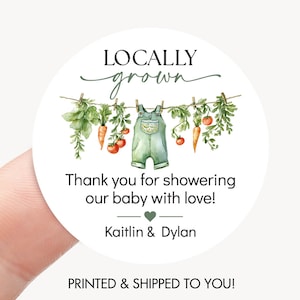 May include: A round sticker with the text "LOCALLY grown" above a watercolor illustration of vegetables and baby overalls hanging on a clothesline. The text "Thank you for showering our baby with love!" is below the illustration, with names and a heart symbol.