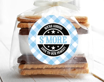 May include: A clear cellophane bag filled with a stack of graham crackers, chocolate squares, and marshmallows. A blue and white gingham patterned circle sticker with black text reads "We're adding S'more love to our family!"