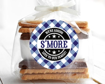 May include: A clear cellophane bag tied with a white ribbon, containing graham crackers, chocolate, and marshmallows. A round sticker with blue and white checkered pattern reads "We're adding S'more Love to our Family!"