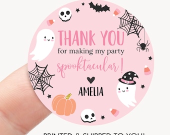 May include: A round pink sticker featuring the text THANK YOU for making my party spooktacular! AMELIA. The design includes illustrations of white ghosts, a pumpkin, skulls, spider webs, a spider, bats, and candy corn pieces.