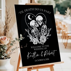 May include: A black wedding welcome sign on a wooden easel featuring white line art of two skeletons embracing inside a heart shape with flowers, stars, and a crescent moon. The text reads 'TIL DEATH DO US PART', 'WELCOME TO THE WEDDING OF', 'Kaitlin & Robert', and 'OCTOBER 31, 2026'.