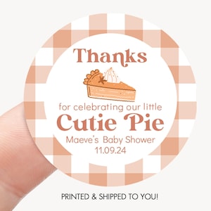 May include: A round sticker with a white and peach gingham pattern border. The center features text that reads "Thanks for celebrating our little Cutie Pie" with a pie graphic. The sticker also includes the text "Maeve's Baby Shower 11.09.24".