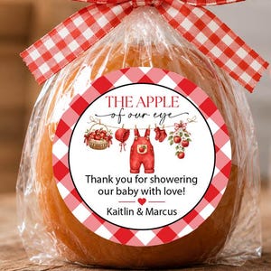 May include: A caramel apple wrapped in clear plastic with a red and white gingham ribbon bow. A round sticker features the text THE APPLE of our eye, Thank you for showering our baby with love! Kaitlin & Marcus, and illustrations of baby clothes.