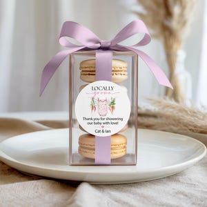 May include: A clear box tied with a lavender ribbon, containing four macarons. A round sticker on the box reads "Locally Grown" and "Thank you for showering our baby with love!" with a baby clothes graphic. The box sits on a white plate.