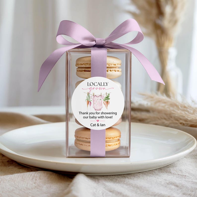 May include: A clear box tied with a lavender ribbon, containing four macarons. A round sticker on the box reads "Locally Grown" and "Thank you for showering our baby with love!" with a baby clothes graphic. The box sits on a white plate.
