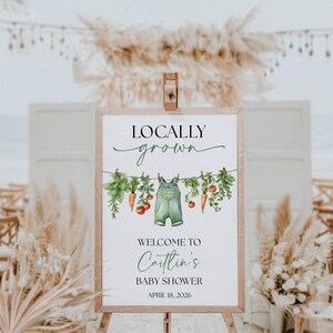 May include: A wooden framed sign with the words "Locally Grown" in a script font. Below is a watercolor illustration of vegetables and overalls hanging on a clothesline. The sign welcomes guests to "Caitlin's Baby Shower" on April 18, 2026.
