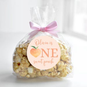 May include: Peach-themed favor bag with a pink ribbon and a round sticker that says "Olivia is one sweet peach". The sticker features a peach illustration.