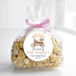 K&ouml;nnte beinhalten: Ein klarer Cellophanbeutel gef&uuml;llt mit Karamell-Popcorn, gebunden mit einem rosa Band. Ein runder Aufkleber zeigt eine Aquarellillustration eines Marktkarrens mit Blumen und dem Text "She's fresh off THE MARKET Claire's Bridal Shower 5.31.25."