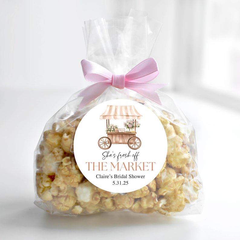 K&ouml;nnte beinhalten: Ein klarer Cellophanbeutel gef&uuml;llt mit Karamell-Popcorn, gebunden mit einem rosa Band. Ein runder Aufkleber zeigt eine Aquarellillustration eines Marktkarrens mit Blumen und dem Text "She's fresh off THE MARKET Claire's Bridal Shower 5.31.25."