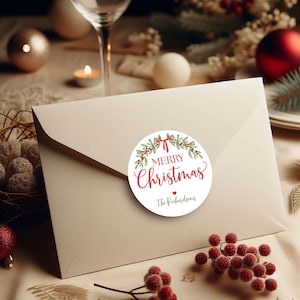 May include: A cream-colored envelope sealed with a round sticker featuring a watercolor wreath of green leaves and red berries. The sticker displays the red text Merry Christmas, a small red heart, and the black text The Richardsons.