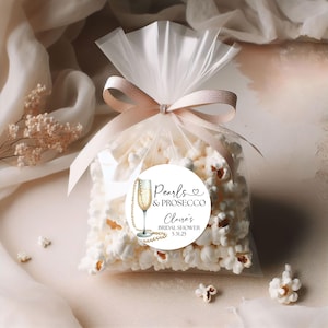May include: White favor bags filled with popcorn and tied with a pink ribbon. A round sticker with a champagne glass, pearls, and the text "Pearls & Prosecco,  Claire's Bridal Shower 5.31.25" is attached to the bag.