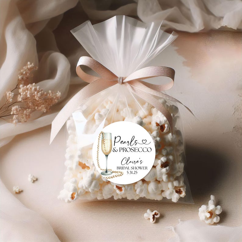 May include: White favor bags filled with popcorn and tied with a pink ribbon. A round sticker with a champagne glass, pearls, and the text "Pearls & Prosecco,  Claire's Bridal Shower 5.31.25" is attached to the bag.