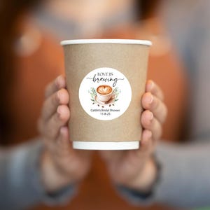 May include: A brown paper coffee cup with a white rim, held by a person. The cup has a circular sticker with the words "Love is Brewing" and a coffee cup design. Below the design is text that reads "Caitlin's Bridal Shower 11-8-25".