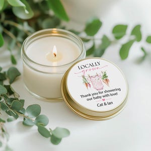 May include: A lit candle in a clear glass jar next to a gold-rimmed lid. The lid features the text "Locally Grown" and "Thank you for showering our baby with love!" with a pink onesie graphic. Eucalyptus leaves surround the candle.