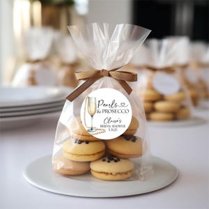 May include: A clear plastic bag filled with small, round, light brown cookies. The bag is tied with a brown ribbon and has a white round sticker with black text that reads "Pearls & Prosecco,  Cloe's Bridal Shower, 5.31.25".
