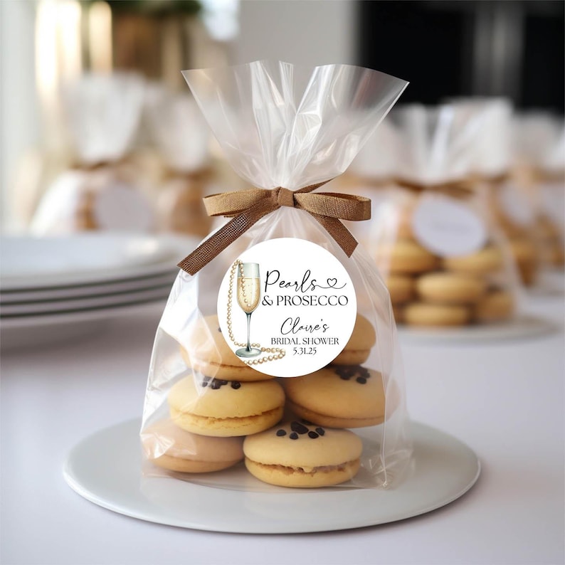 May include: A clear plastic bag filled with small, round, light brown cookies. The bag is tied with a brown ribbon and has a white round sticker with black text that reads "Pearls & Prosecco,  Cloe's Bridal Shower, 5.31.25".