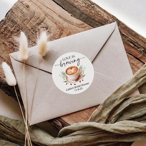 May include: A round sticker with the text "Love is Brewing" and a coffee cup design. The sticker is on a light brown envelope, with dried flowers and a green ribbon. The sticker also includes the text "Caitlin's Bridal Shower 11-8-25".
