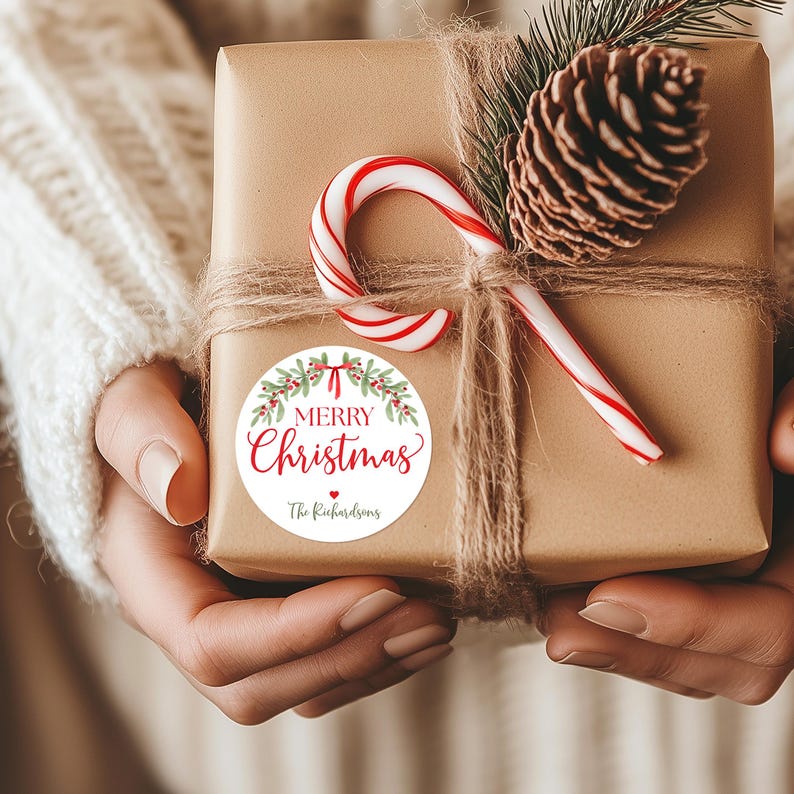 May include: A round white sticker with a green mistletoe wreath illustration, red text reading Merry Christmas, and the name The Richardsons below a small red heart, attached to a brown gift box wrapped in twine with a candy cane.