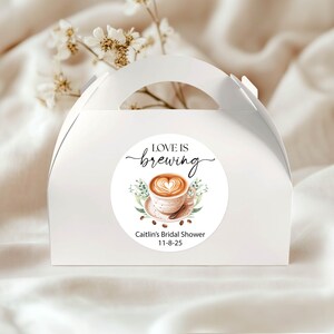 May include: A white favor box with a handle, featuring a circular sticker. The sticker has the text "LOVE IS brewing" above a coffee cup design with a heart-shaped foam. Below the coffee cup is the text "Caitlin's Bridal Shower 11-8-25".
