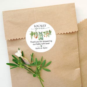 May include: A round sticker with the text "Locally Grown" and a watercolor illustration of vegetables and overalls. The sticker reads "Thank you for showering our baby with love!" and includes the names "Kaitlin & Dylan". The sticker is on a brown paper bag.