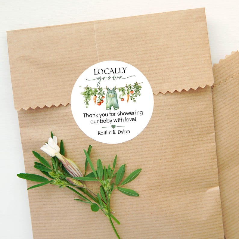 May include: A round sticker with the text "Locally Grown" and a watercolor illustration of vegetables and overalls. The sticker reads "Thank you for showering our baby with love!" and includes the names "Kaitlin & Dylan". The sticker is on a brown paper bag.