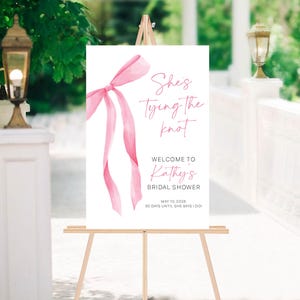 以下が含まれることがあります： ピンクの水彩画のリボンと「She's tying the knot. Welcome to Kathy's Bridal Shower. May 10, 2025. 30 days until she says I do!」というテキストが書かれた白い看板