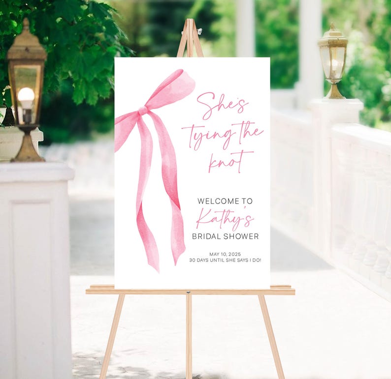 以下が含まれることがあります： ピンクの水彩画のリボンと「She's tying the knot. Welcome to Kathy's Bridal Shower. May 10, 2025. 30 days until she says I do!」というテキストが書かれた白い看板
