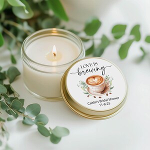 May include: A lit, white candle in a clear glass jar next to a gold-rimmed lid. The lid features the text "LOVE IS brewing" with a coffee cup design and the text "Caitlin's Bridal Shower 11-8-25".