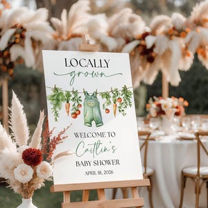 May include: A white sign on a wooden easel with the text "Locally Grown" and "Welcome to Caitlin's Baby Shower April 18, 2026." The sign features watercolor illustrations of vegetables and overalls.