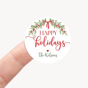 May include: A round white sticker featuring a watercolor illustration of green holly leaves and red berries tied with a red ribbon. The text reads "HAPPY holidays" in red and "The Wilsons" in dark green, with a small red heart in between.