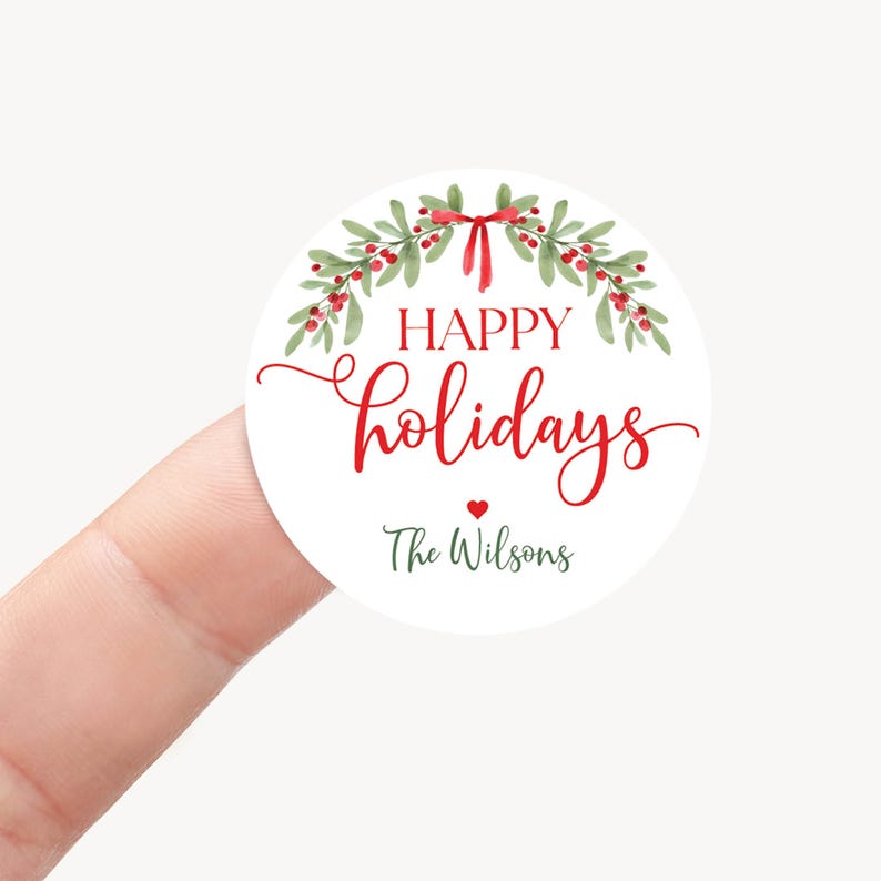 May include: A round white sticker featuring a watercolor illustration of green holly leaves and red berries tied with a red ribbon. The text reads "HAPPY holidays" in red and "The Wilsons" in dark green, with a small red heart in between.