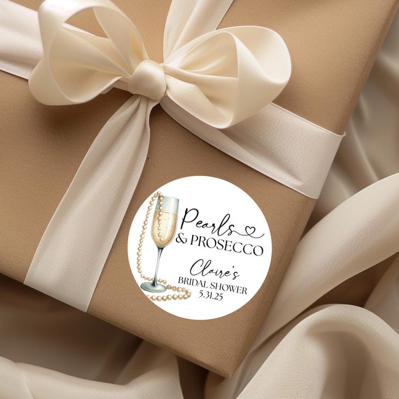 May include: A white round sticker with a black and white illustration of a champagne flute with a pearl necklace around it. The text on the sticker reads "Pearls & Prosecco Claire's Bridal Shower 5.31.25".