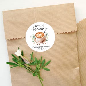 May include: A round sticker with the text "Love is brewing" in elegant script, featuring a watercolor illustration of a coffee cup with a heart-shaped design. The sticker also includes the text "Caitlin's Bridal Shower 11-8-25". The sticker is placed on a brown paper bag.