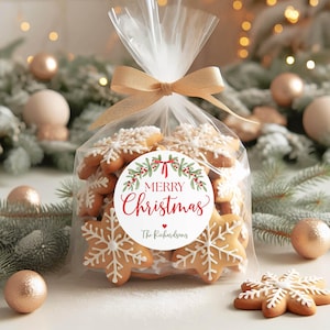 May include: A clear cellophane bag filled with snowflake-shaped gingerbread cookies, tied with a gold ribbon. A round white label on the front features a green holly wreath, red berries, and the text Merry Christmas and The Richardsons.