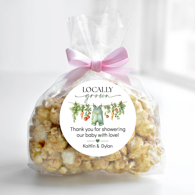 May include: A clear cellophane bag filled with caramel popcorn, tied with a pink ribbon. A round sticker on the bag reads "Locally Grown" and "Thank you for showering our baby with love!" with a watercolor illustration of baby clothes and vegetables.