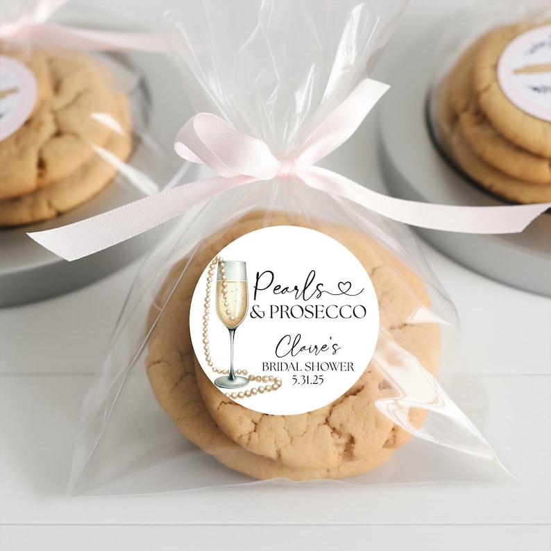 May include: A white round sticker with black text that reads "Pearls & Prosecco Claire's Bridal Shower 5.31.25". The sticker features a gold champagne flute with a pearl necklace wrapped around it. The sticker is attached to a clear plastic bag with a pink ribbon.