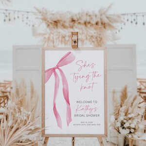 以下が含まれることがあります： 白い背景に「She's tying the knot」と「Welcome to Kathy's Bridal Shower」のテキストが書かれた、ピンクの水彩画の弓。日付「May 10, 2025」とテキスト「30 days until she says I do!」も含まれています。