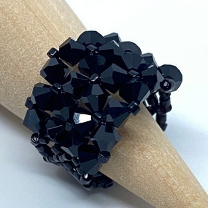 May include: A black beaded ring with a geometric design. The ring is made with black crystal beads and is on a wooden ring mandrel.
