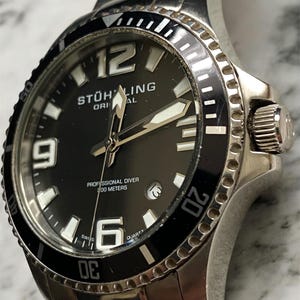 May include: A stainless steel Stührling Original quartz watch with a black dial. The watch has large white numerals at the 12, 9, and 6 o'clock positions, and a date window. The black bezel has white markings, and the watch is water resistant to 100 metres.