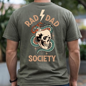 Peut inclure: T-shirt vert olive avec un motif au dos. Le motif représente un crâne entouré d'un serpent, avec les mots "RAD DAD SOCIETY" et un éclair. Le serpent est vert et rouge, et le crâne est blanc.