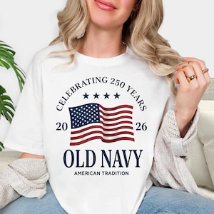 May include: A white t-shirt featuring a graphic of a waving American flag with four blue stars above it. The text reads CELEBRATING 250 YEARS, 20 26, OLD NAVY, and AMERICAN TRADITION in navy blue lettering centred on the front of the garment.