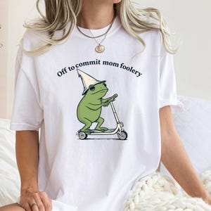 May include: White t-shirt with the text "Off to commit mom foolery." The shirt features a green frog wearing a pointed hat and riding a scooter. The frog is the central graphic element.