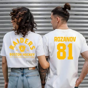 May include: White t-shirts with yellow lettering. One shirt reads "RAIDERS BOSTON HOCKEY EST. NINETEEN TWENTY FOUR" on the back. The other shirt displays "ROZANOV 81". The shirts are worn by two people.