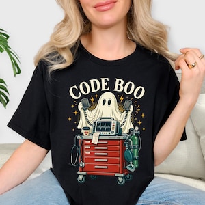 May include: A black t-shirt featuring a graphic of a white ghost holding defibrillator paddles above a red medical crash cart. The cart includes a heart monitor, oxygen tank, and stethoscope. The text CODE BOO is printed in cream above the ghost.