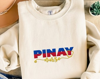 May include: A cream-colored sweatshirt with the text "PINAY NURSE" in red, blue, and gold letters. The letters are stylized to look like the Philippine flag. A gold stethoscope is next to the text.