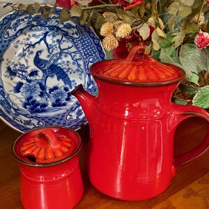 May include: A red enamel teapot and sugar bowl set with a matching lid. The teapot has a curved handle and spout. A blue and white porcelain plate with a peacock design is in the background.