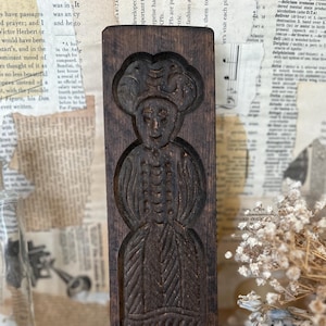 May include: A vintage, dark brown wooden cookie mold featuring a carved figure of a woman in a bonnet and dress. The rectangular mold is intricately detailed with a folk art style, showcasing the woman's features and clothing.