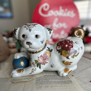May include: A white ceramic dog figurine with floral and butterfly designs. The dog is lying down, holding a blue and gold ball. The figurine has a gold-colored collar and tail. The background includes a red sign that says "Cookies for Santa".