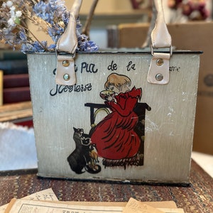 May include: Vintage metal tote bag with a light beige finish, featuring a charming illustration of a girl in a red dress with a black cat. The bag has beige leather handles and accents. The text on the bag is in French.