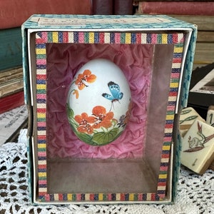 May include: A vintage, white egg-shaped ornament with orange floral and blue butterfly designs, presented in a decorative box. The box has a colorful striped border and a pink, tissue-like interior. The egg is a unique decorative item.